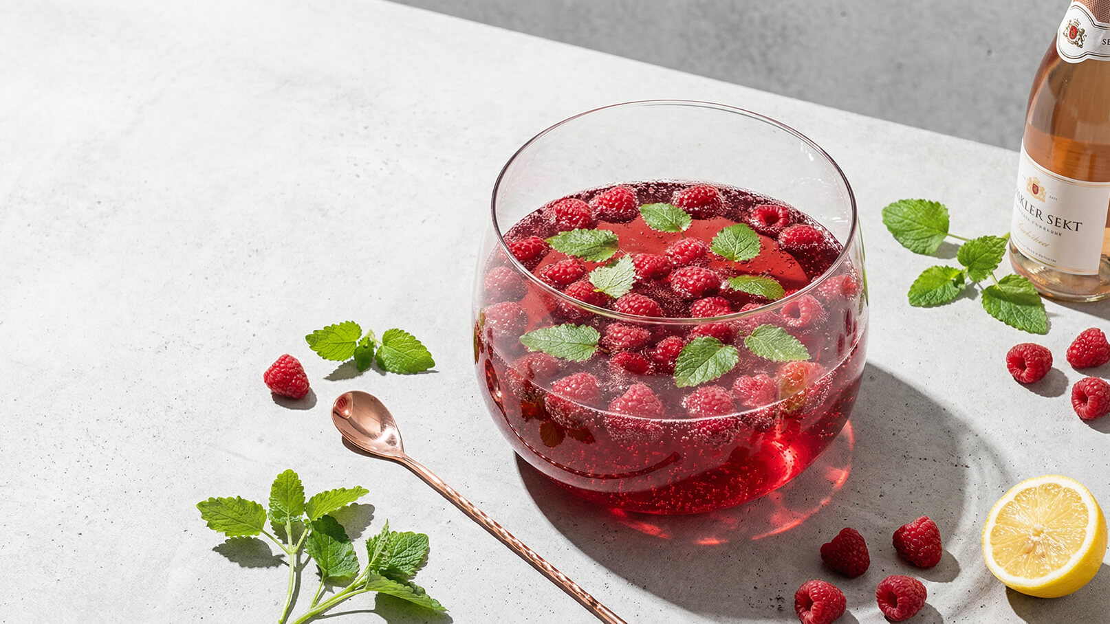 Eine große Glasschüssel enthält sprudelnden roten Sekt mit frischen Himbeeren und Minzblättern. Neben der Schüssel liegen einige Himbeeren, Minzblätter und eine halbe Zitrone auf einer grauen Oberfläche. Ein Kupferlöffel ergänzt die Anordnung.