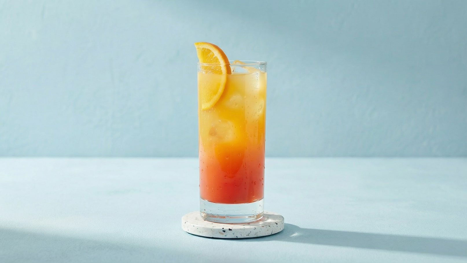 Ein hochstehendes Glas mit einem bunten Getränk, das von orange über gelb bis rot verläuft. Es enthält Eiswürfel und ist mit einer Zitronenscheibe garniert. Das Glas steht auf einem weißen Untersetzer vor einer hellblauen Wand.
