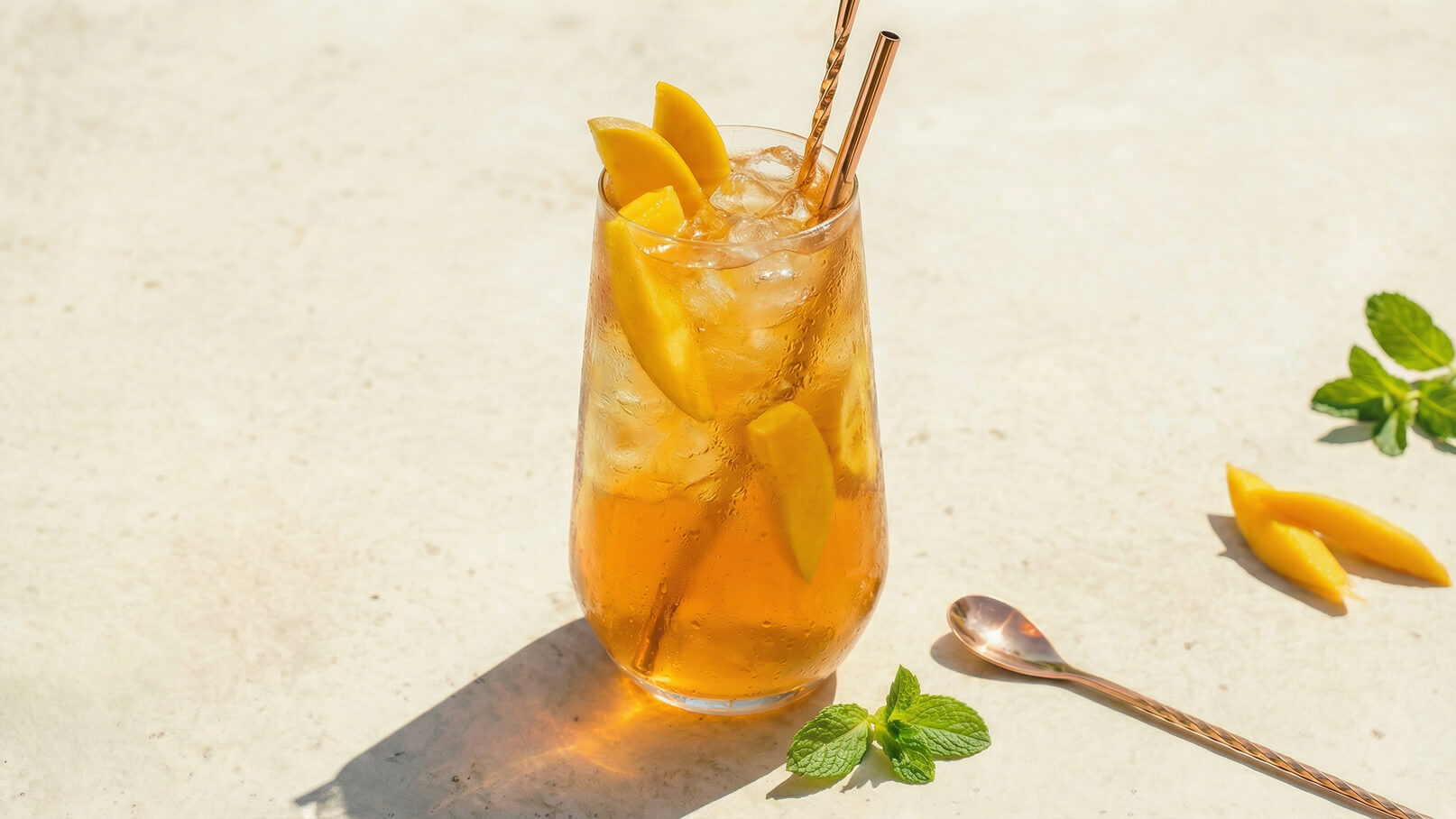 Ein Glas mit einem erfrischenden Getränk, gefüllt mit Eiswürfeln und frischen Mango-Stücken, steht auf einer hellen Oberfläche. Zwei Gold-Getränkehalme stecken im Glas, und einige Minzblätter sowie Mangostücke liegen daneben.