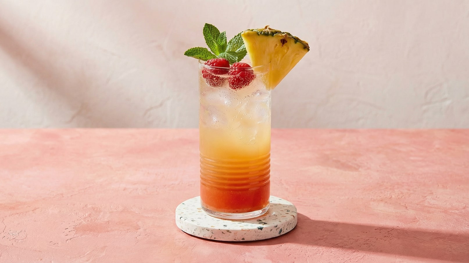 Ein erfrischender Cocktail in einem langen Glas, gefüllt mit Eis und einer bunten Flüssigkeit. Obenauf befinden sich frische Himbeeren, ein Ananasstück und Minzblätter als Dekoration. Das Glas steht auf einem dekorativen Untersetzer auf einem rosa Tisch.