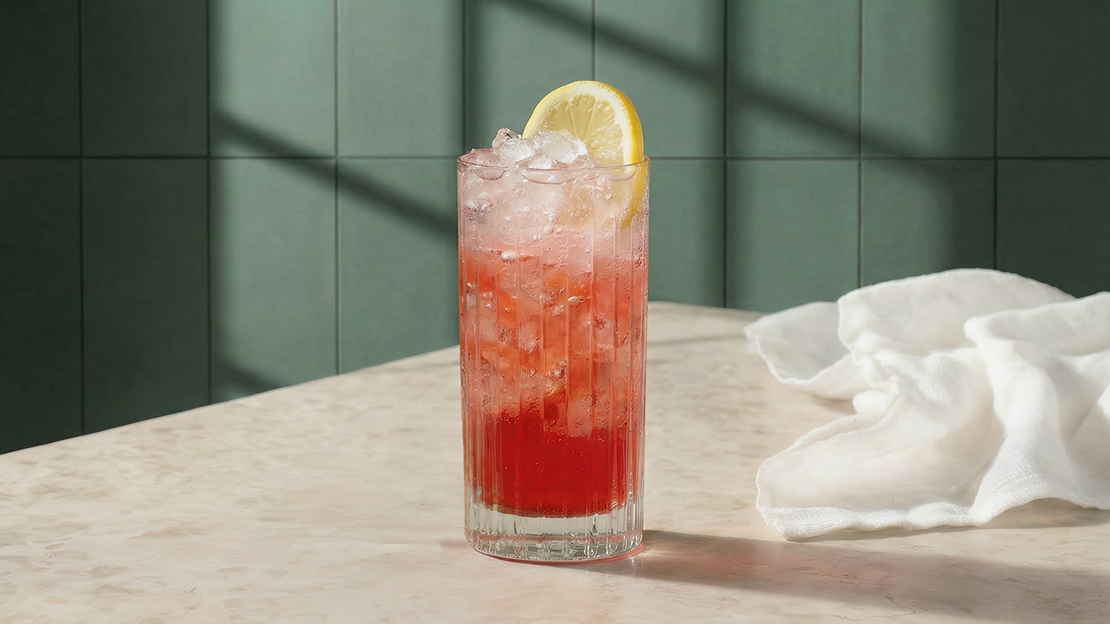 Ein erfrischendes Getränk in einem hohen Glas, gefüllt mit Eis und einer pinken Flüssigkeit. Obenauf liegt eine Zitronenscheibe. Im Hintergrund sind grüne Wände und ein Tisch aus hellem Material sichtbar.