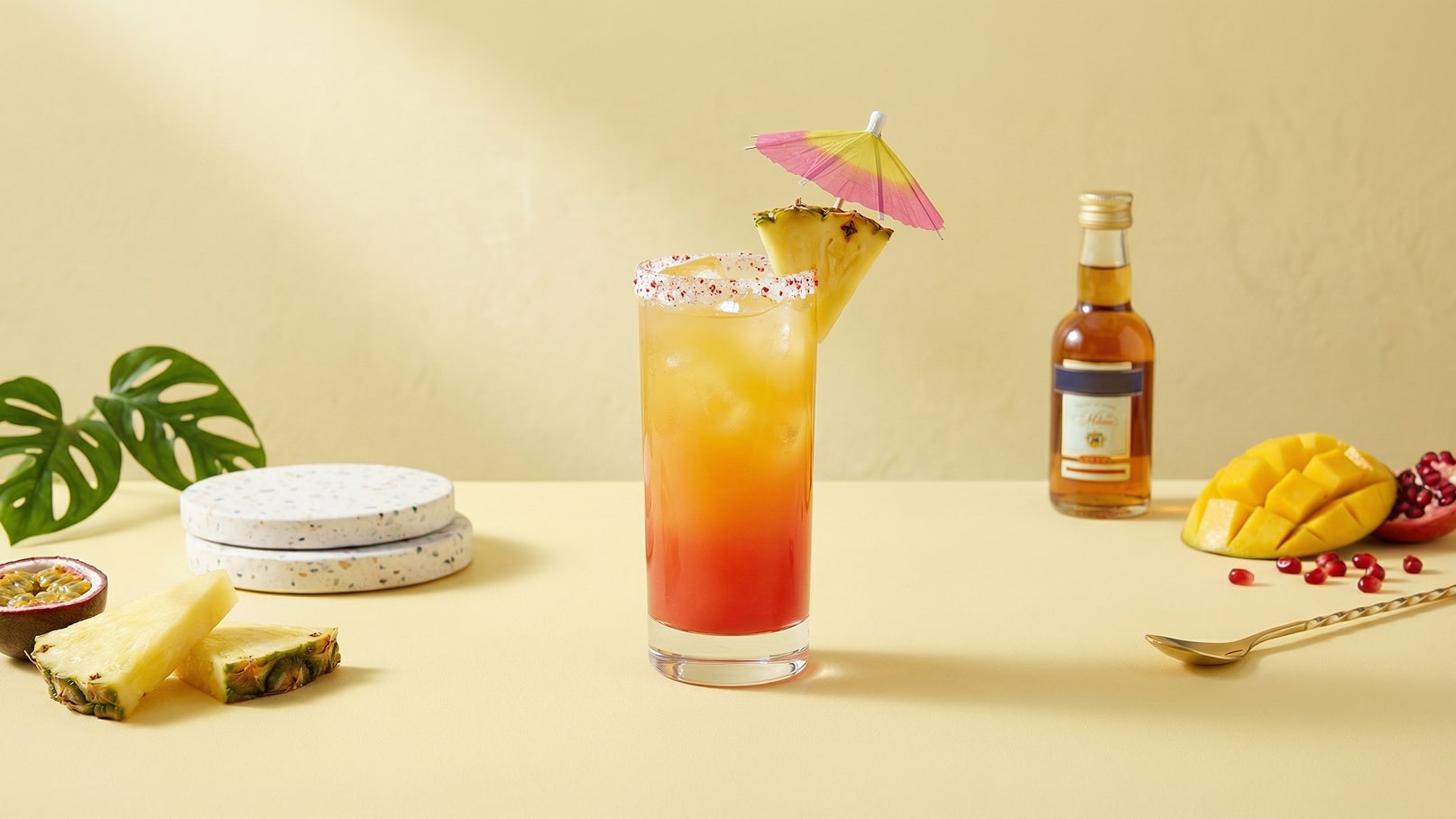 Ein bunter Cocktail in einem hohen Glas, dekoriert mit einem kleinen Regenschirm und einer Ananasscheibe. Im Hintergrund sind geschnittene Früchte wie Mango und Granatapfel sowie eine Flasche Rum und einige Untersetzer zu sehen. Der Hintergrund ist gelb.