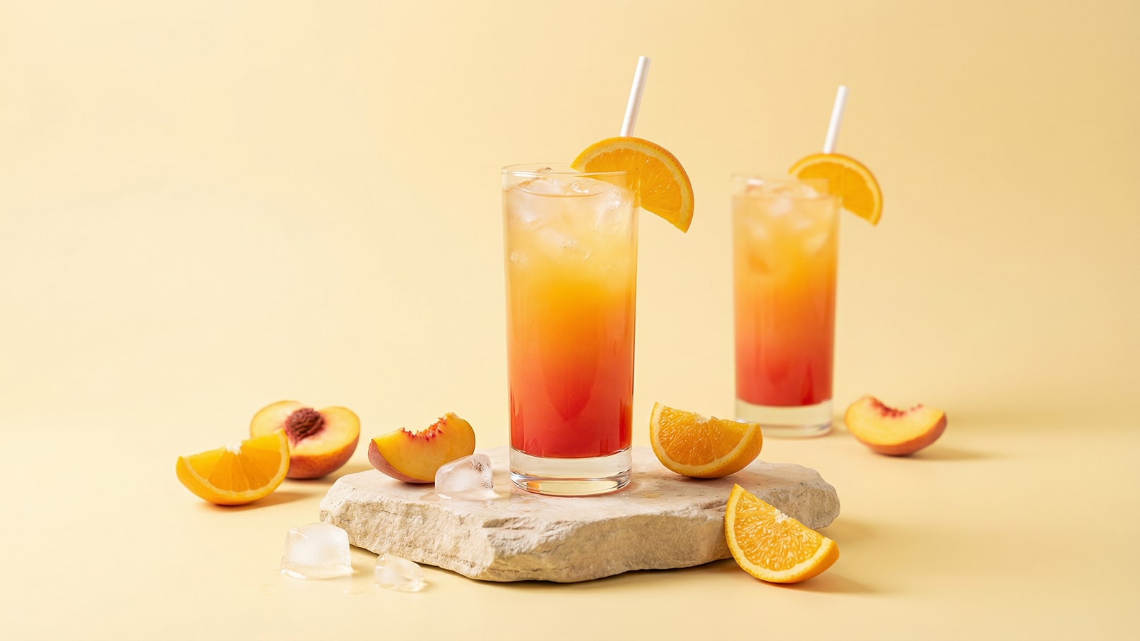 Zwei hohe Gläser mit einem bunten Getränk aus Eiswürfeln, Orange und einem Pfirsichstück dekoriert. Rundherum liegen frische Orangenscheiben und Pfirsichhälften auf einem hellen Untergrund. Die Atmosphäre wirkt sommerlich und einladend.
