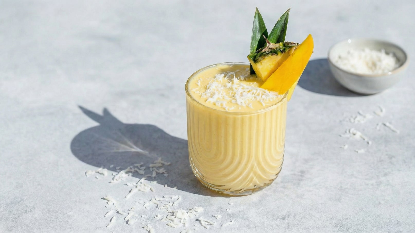 Ein stylisches Glas mit einem cremigen, hellgelben Smoothie, garniert mit einem Stück Ananas und einigen Kokosraspeln obenauf. Im Hintergrund ist eine kleine Schüssel mit weiteren Kokosraspeln zu sehen, auf einem hellen, neutralen Untergrund.