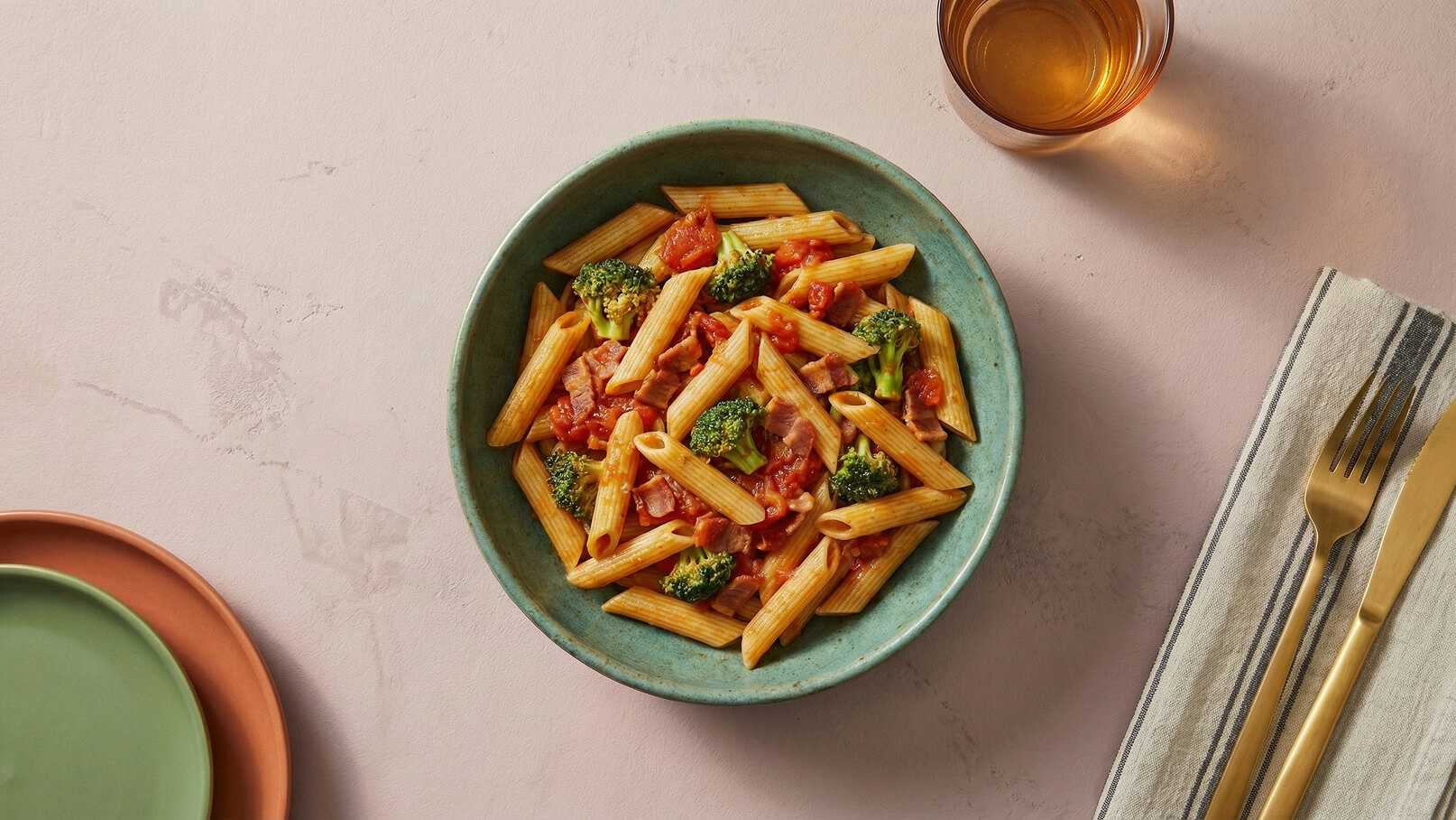 Eine große grüne Schüssel enthält Penne-Pasta, überzogen mit einer Tomatensauce und Brokkoliröschen. Daneben steht ein Glas mit Getränk und Besteck auf einem gestreiften Serviette. Der Hintergrund ist in sanften, neutralen Farben gehalten.