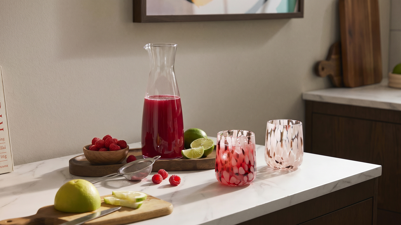 Auf einer Marmortheke stehen eine Karaffe mit rotem Getränk, zwei Gläser mit Eis und Limettenscheiben, frische Himbeeren und ein paar Limettenhälften auf einem Holzbrett. Ein frischer, einladender Anblick einer sommerlichen Getränkeszubereitung.