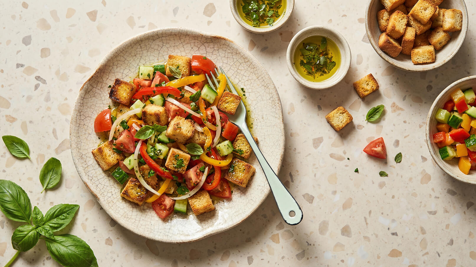 Ein Teller mit einem bunten Salat aus Tomaten, Gurken, Paprika und Zwiebeln, garniert mit frischen Kräutern und gerösteten Brotwürfeln. Daneben befinden sich Schalen mit Kräuteröl und zusätzlichen Brotwürfeln. Der Hintergrund ist ein helles, strukturiertes Material.