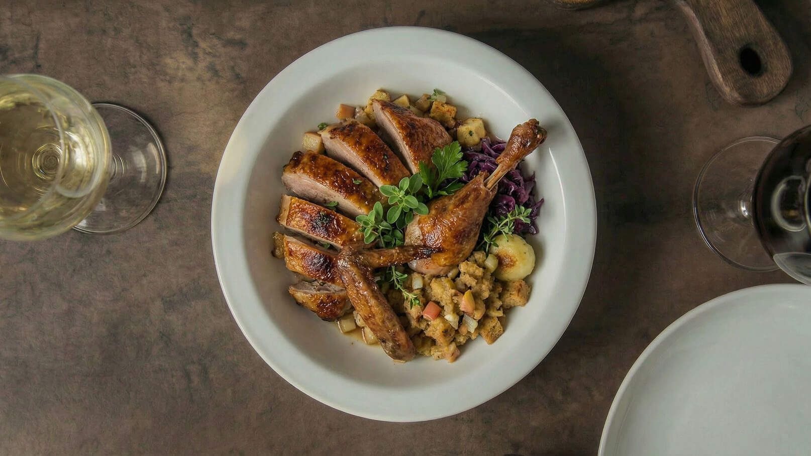 Ein Teller mit gebratenem Geflügel, aufgeschnittene Stücke, zwei Keulen und eine Portion Füllung aus Brot und Äpfeln. Daneben liegt ein kleiner Haufen roter Kohl. Im Hintergrund sind Gläser für Wein und Wasser zu sehen.