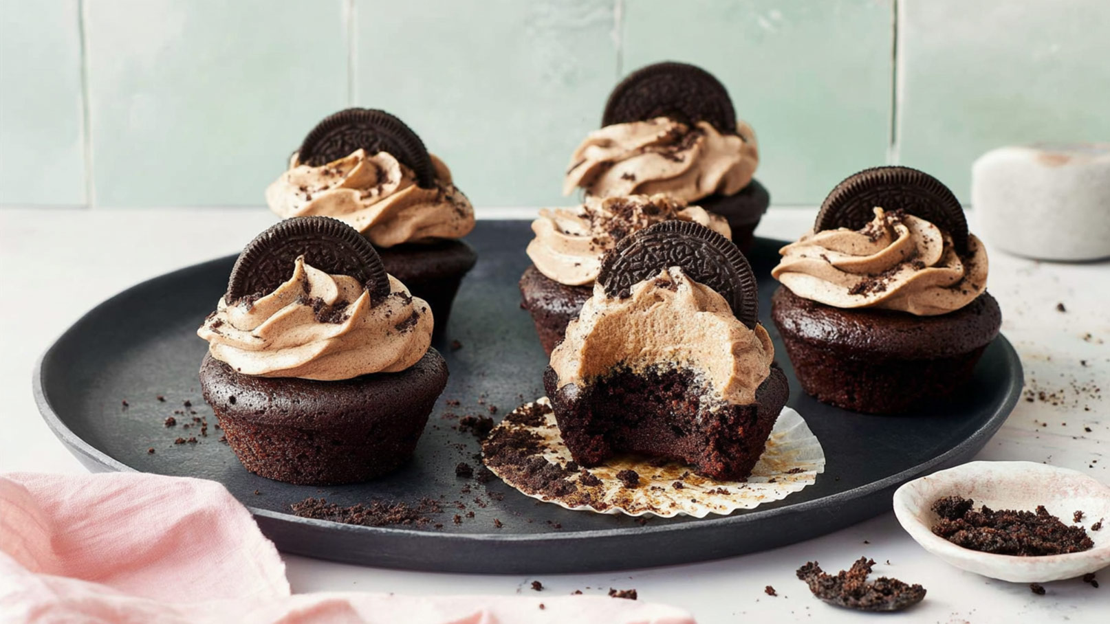 Ein schwarzer Teller mit fünf Schokoladen-Cupcakes, die mit einer cremigen Karamell-Buttercreme und einem Oreo-Keks dekoriert sind. Einer der Cupcakes hat einen Biss, wobei die schokoladige Füllung sichtbar ist. Die Oberfläche ist mit Keksbröseln verziert.