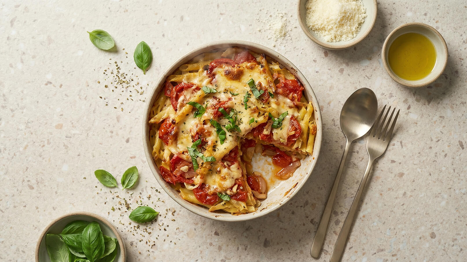 Eine Schüssel mit heißem, gebackenem Pasta-Gericht, belegte mit geschmolzenem Käse, Tomaten und frischen Kräutern. Neben der Schüssel sind einige frische Basilikumblätter und kleine Schalen mit Olivenöl und geriebenem Käse platziert.