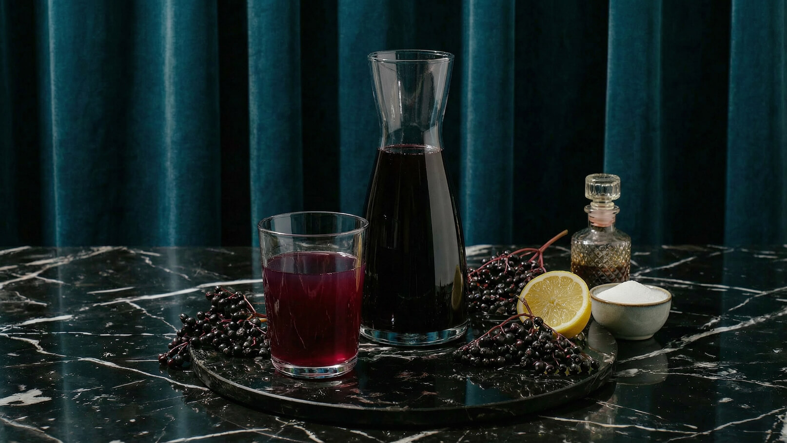 Auf einem dunklen Marmor-Tisch steht eine Glaskaraffe mit dunklem Saft und ein Glas mit der gleichen Flüssigkeit. Umgeben sind sie von Holunderbeeren, einer aufgeschnittenen Zitrone, einer Zuckerschale und einem kleinen Fläschchen. Der Hintergrund ist in einem satten Blau gehalten.