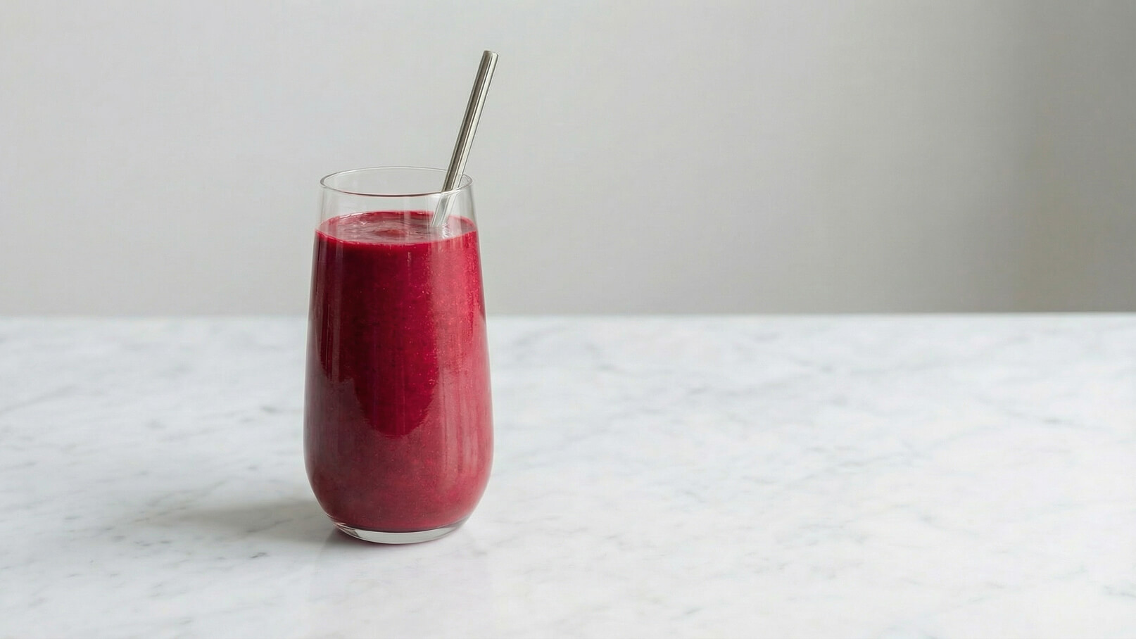 Ein transparentes Glas mit einem leuchtend roten Smoothie steht auf einer marmorierten Oberfläche. Ein Metallstrohhalm ragt aus dem Glas. Der Hintergrund ist neutral und hebt die Farben des Smoothies hervor.