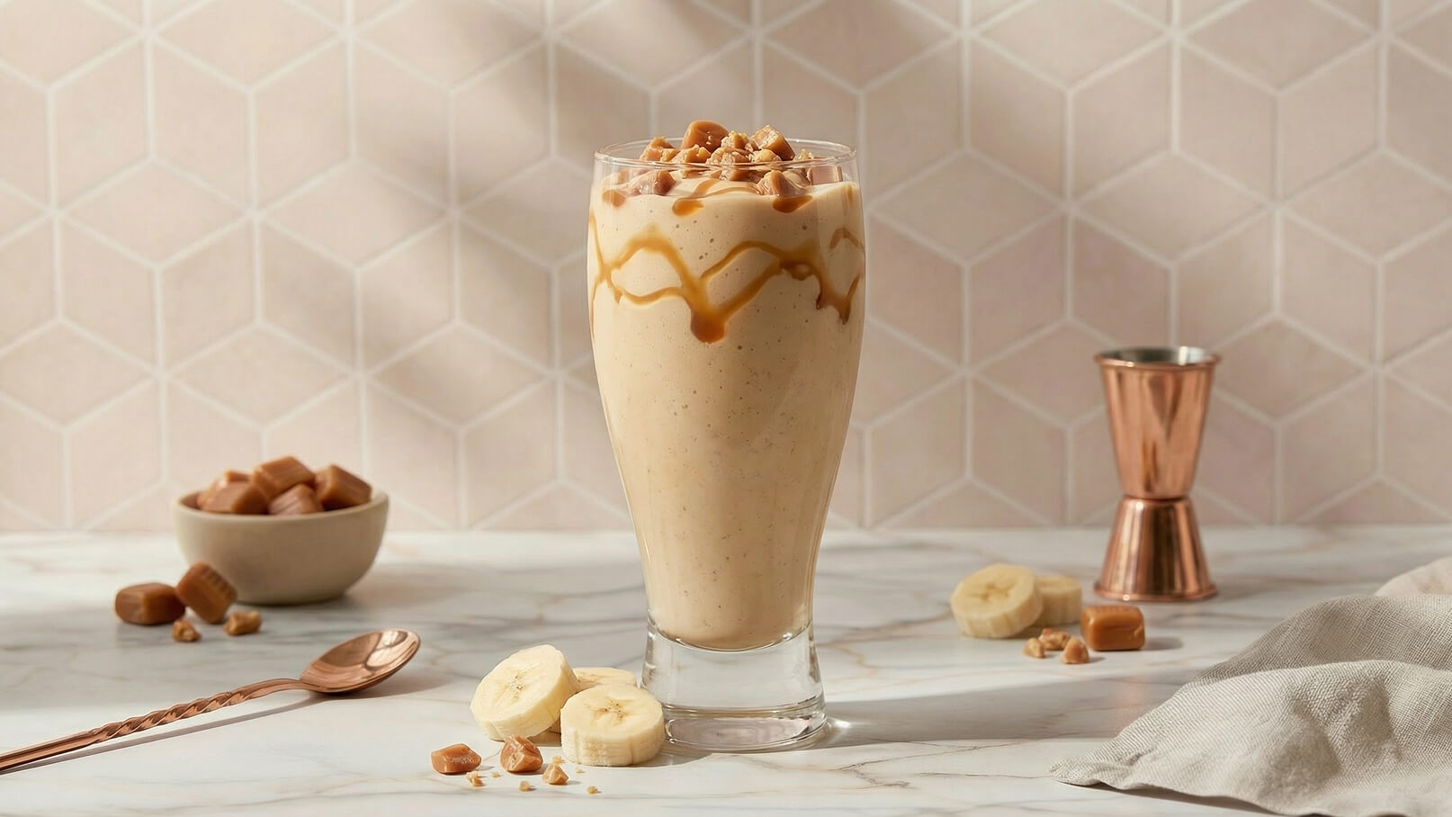 Ein hochgläserner Milchshake mit einer cremigen Konsistenz, garniert mit Karamellsoße und Stückchen. Neben dem Glas liegen einige eingezuckerte Bananenscheiben und Karamellbonbons auf einem marmorierten Tisch. Der Hintergrund ist mit pastellfarbenen geometrischen Mustern dekoriert.