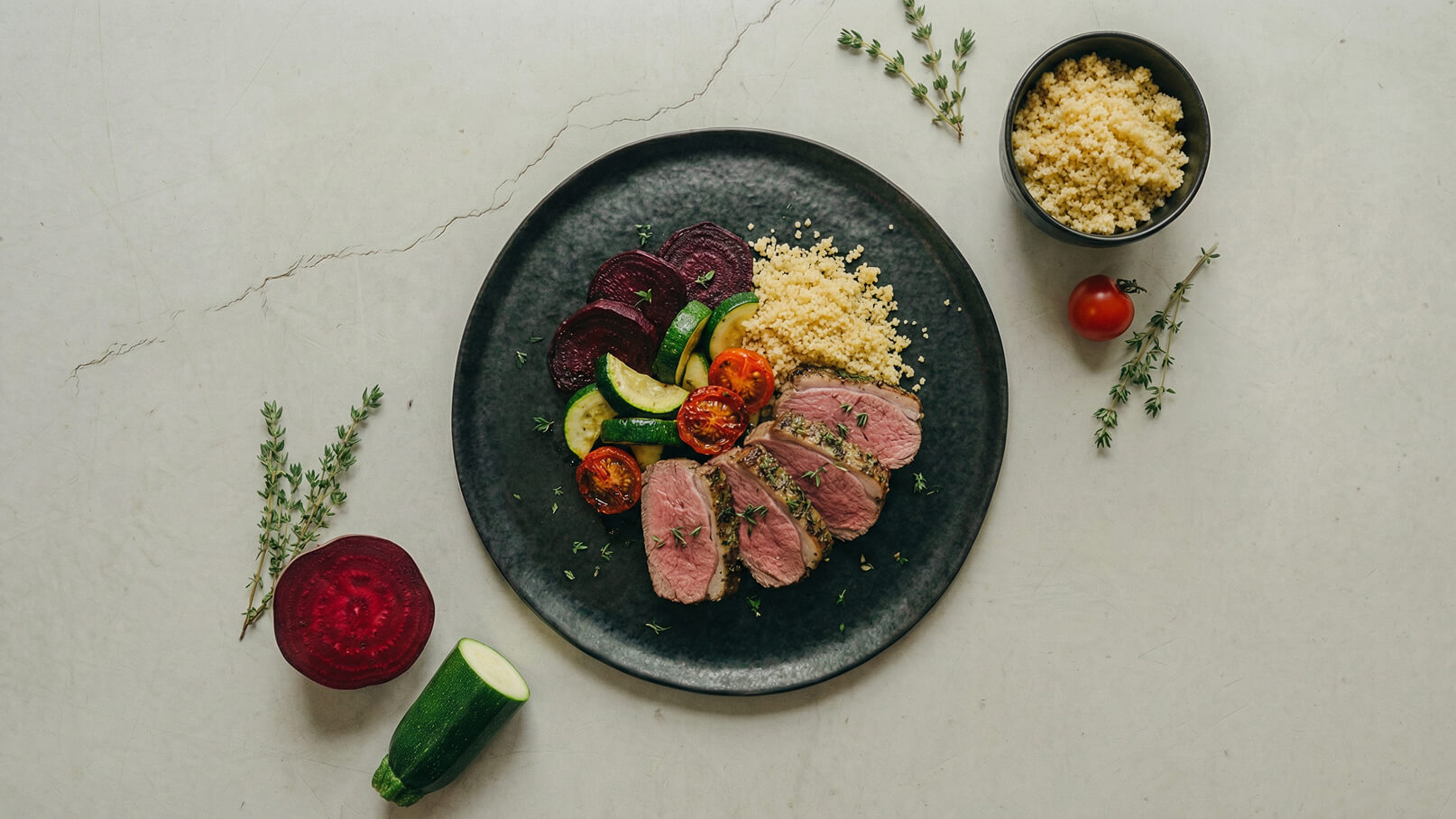Ein schwarzer Teller mit gefülltem Couscous, zarten Scheiben von gebratenem Fleisch, gebratenen Zucchini und Kirschtomaten sowie roter Rübe. Frische Kräuter sind dekorativ verteilt. Der Hintergrund ist hell und einfarbig.