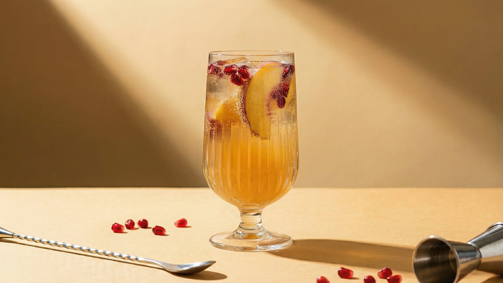Ein transparentes Glas mit einem prickelnden Getränk, das Pfirsichscheiben und Granatapfelkerne enthält. Das Glas steht auf einer flachen, beige Oberfläche, während Schatten eine warme Atmosphäre schaffen. Einige Granatapfelkerne und ein Rührstab sind um das Glas verteilt.
