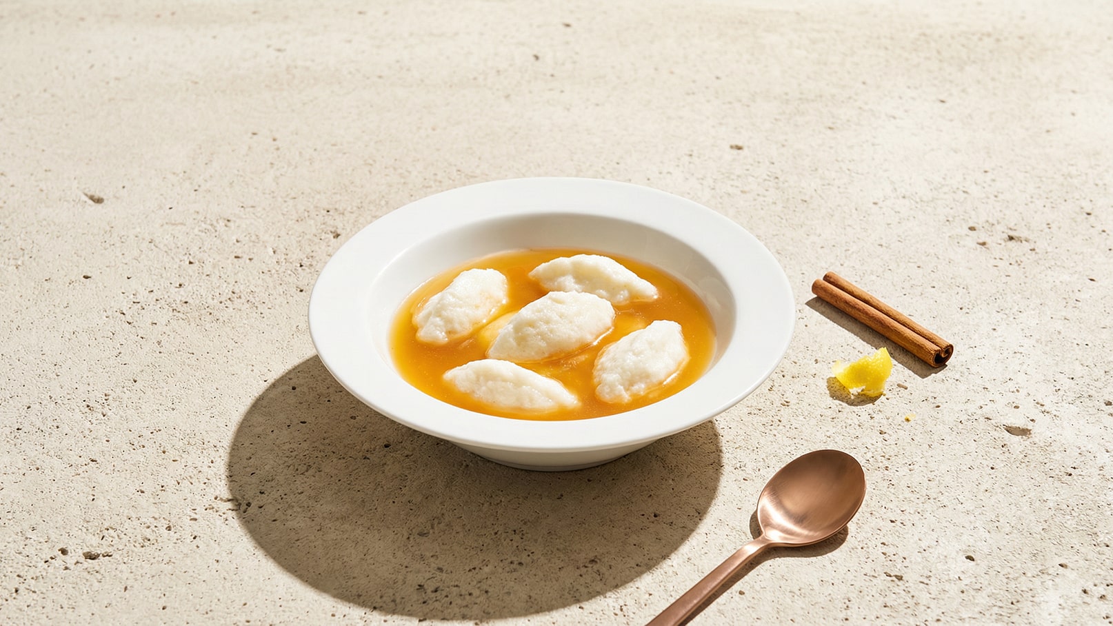 Eine Schüssel mit einer goldenen Brühe, in der mehrere kleine, weiße Klöße schwimmen. Neben der Schüssel liegen ein löffel aus Kupfer und einige Zimtstangen sowie ein Stück Zitronenschale. Der Untergrund ist hell und strukturiert.