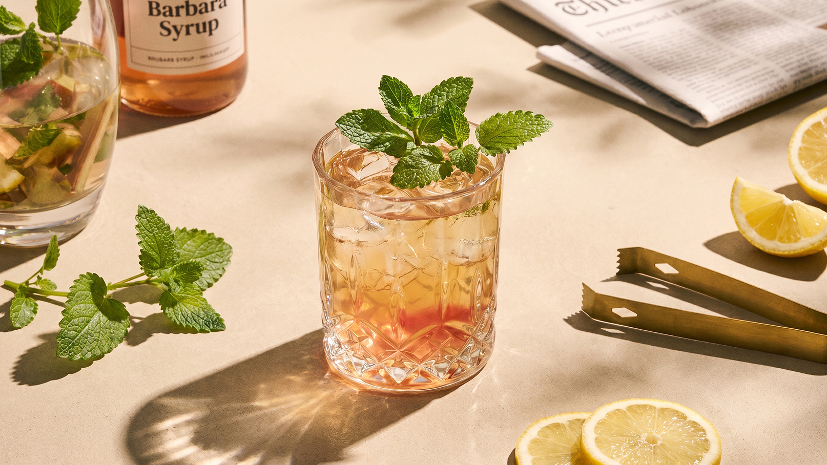 Ein Glas mit einem erfrischenden Getränk, dekoriert mit frischen Minzblättern, steht auf einer hellen Oberfläche. Im Hintergrund sind weitere Zutaten zu sehen, darunter ein Syrup und Zitronenscheiben. Das Bild vermittelt eine einladende, sommerliche Atmosphäre.