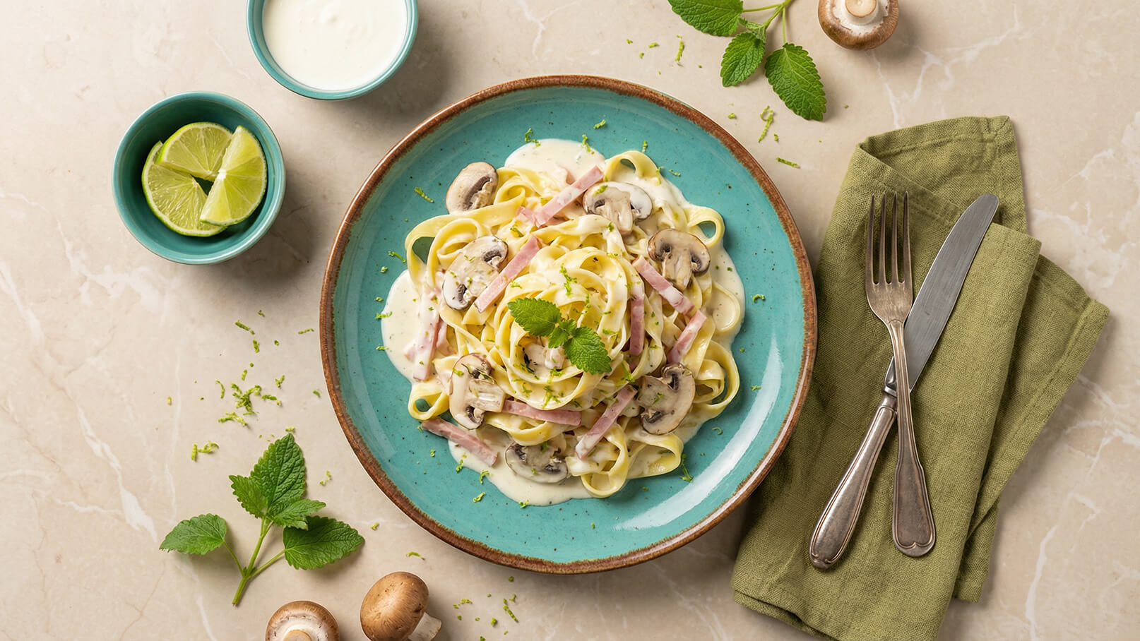 Ein Teller mit Fettuccine in einer cremigen Sauce, garniert mit Champignons und Schinkenstreifen. Frische Minze und Limettenscheiben verleihen dem Gericht Geschmack. Dazu liegen Besteck und ein grünes Serviette.