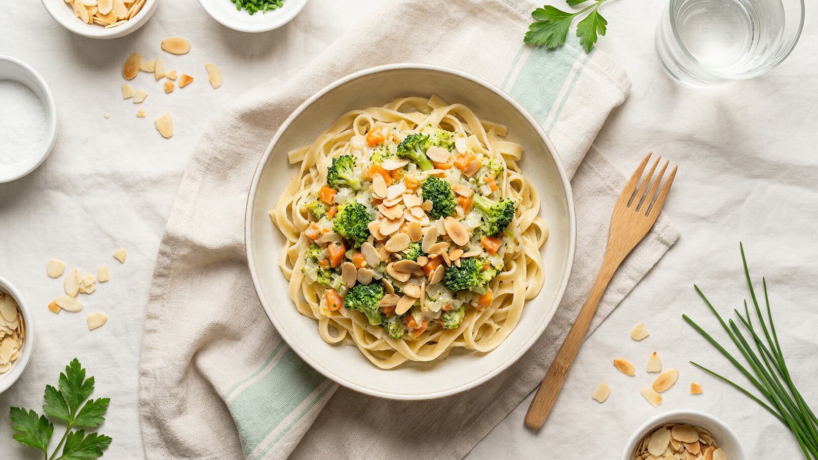 Ein Teller mit Bandnudeln, garniert mit Brokkoli, Karotten und Mandeln. Die Nudeln sind mit einer cremigen Sauce überzogen. Neben dem Teller befinden sich kleine Schalen mit gehobelten Mandeln und frischen Kräutern. Der Hintergrund ist schlicht und in neutralen Farben gehalten.