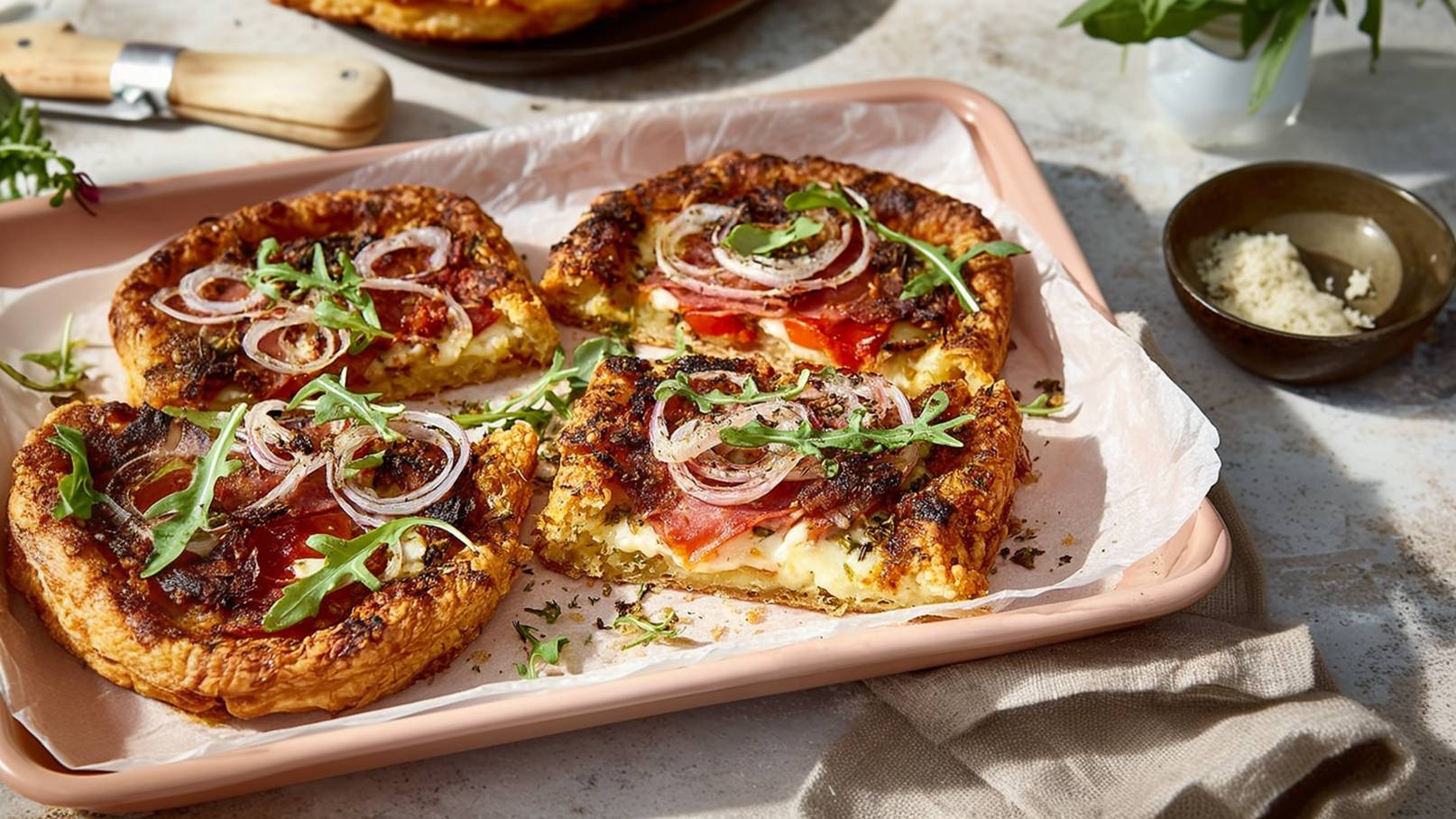 Ein rechteckiger Blech voller gebackener Quiche-Stücke befindet sich auf einem hellen Untergrund. Die Quiche ist mit Schinken, Tomaten, Zwiebeln und frischem Rucola garniert. Neben dem Blech liegt eine kleine Schüssel mit Käse und etwas frischem Grün.