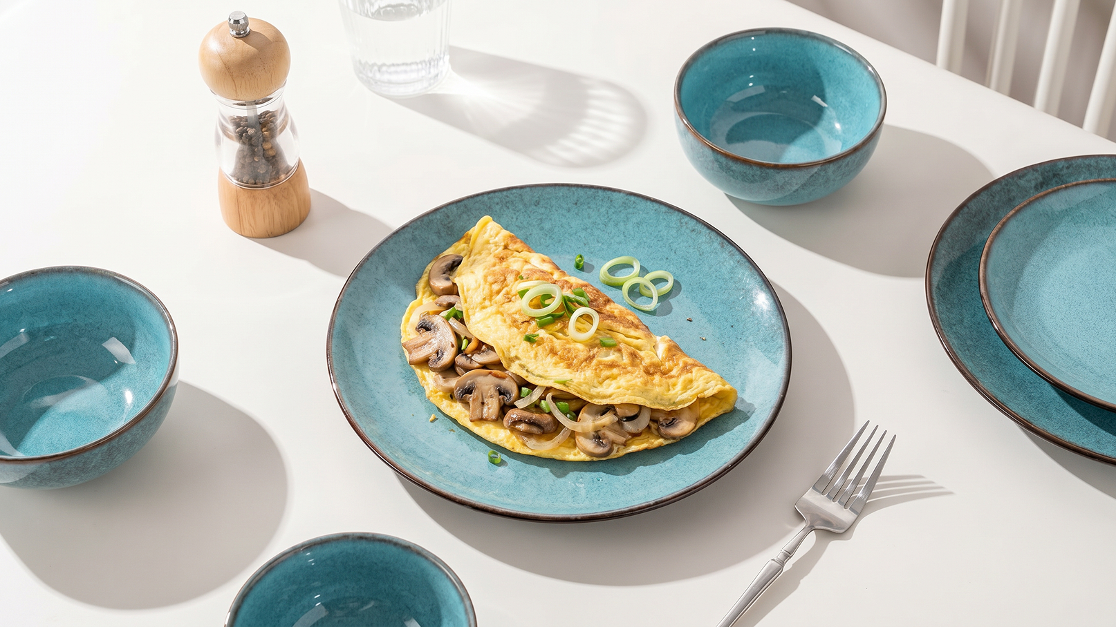 Ein frisches Omelett mit Champignons und Frühlingszwiebeln liegt in einer flachen, blauen Schüssel auf einem gedeckten Tisch. Rundherum sind leere, passende Schüsseln und ein Salzstreuer platziert. Der Tisch hat eine helle, einladende Atmosphäre.