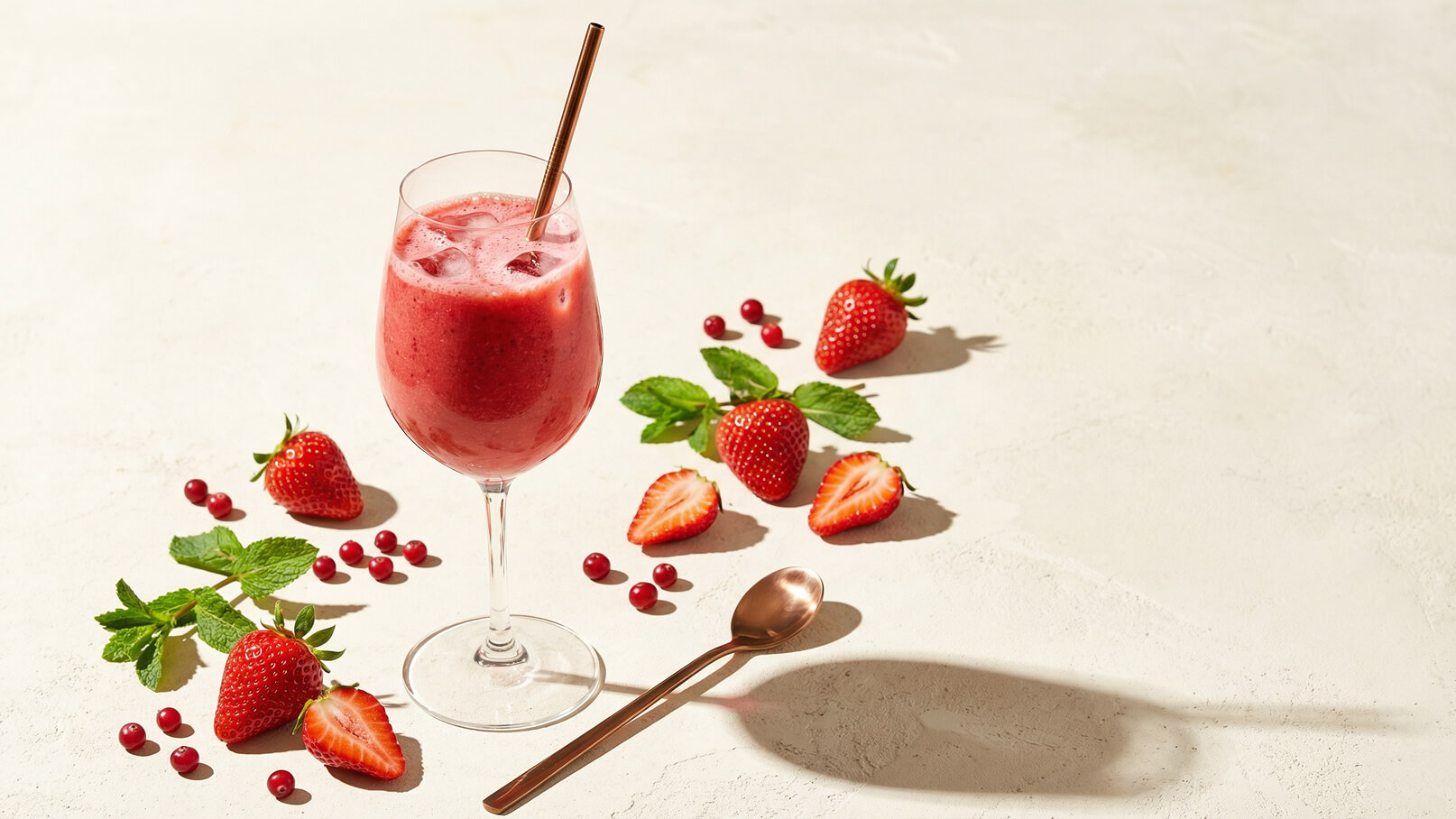 Ein hochglassiger Fruchtshake in einem Kelch, gefüllt mit einer rosa Mischung und Eiswürfeln, steht auf einer hellen Oberfläche. Um ihn herum sind frische Erdbeeren, einige geschnitten, und rote Beeren sowie einige Minzblätter positioniert. Ein Löffel liegt daneben.