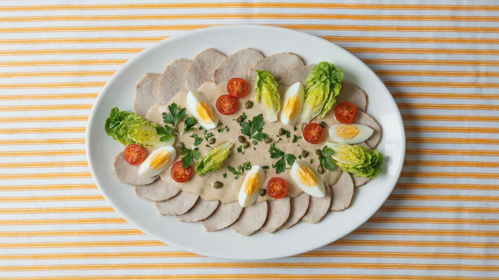 Ein ovaler Teller mit dünn geschnittenem Fleisch, garniert mit halbierten Kirschtomaten, gekochten Eiern und Salatblättern. In der Mitte befindet sich eine cremige Sauce, die mit grünen Kapern und frischer Petersilie dekoriert ist. Der Teller ist auf einem gestreiften Tischcloth platziert.