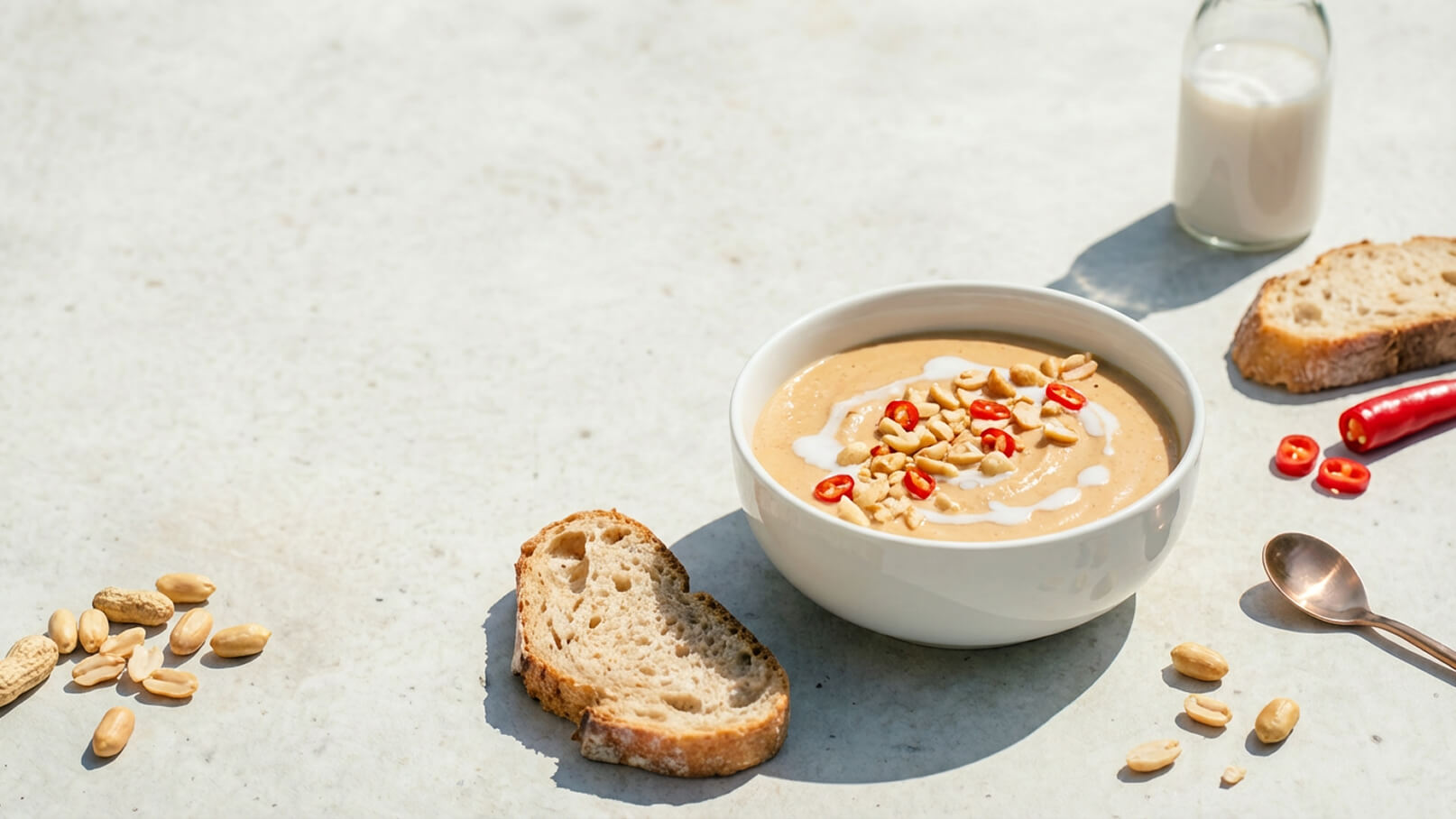 Eine Schüssel mit cremiger Erdnusssuppe, garniert mit roten Chilischoten und gerösteten Erdnüssen. Daneben liegen Scheiben von Brot und einige Erdnüsse auf einer hellen Oberfläche. Im Hintergrund steht ein Glas mit Milch.