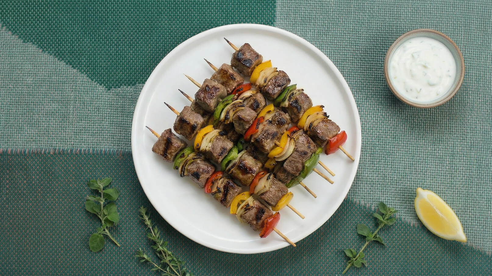 Auf einem weißen Teller liegen gegrillte Spieße mit zarten Fleischstücken und buntem Gemüse, darunter Paprika und Zwiebeln. Neben dem Teller befindet sich eine kleine Schüssel mit einer Joghurtsauce und eine halbe Zitrone. Der Hintergrund ist grün.