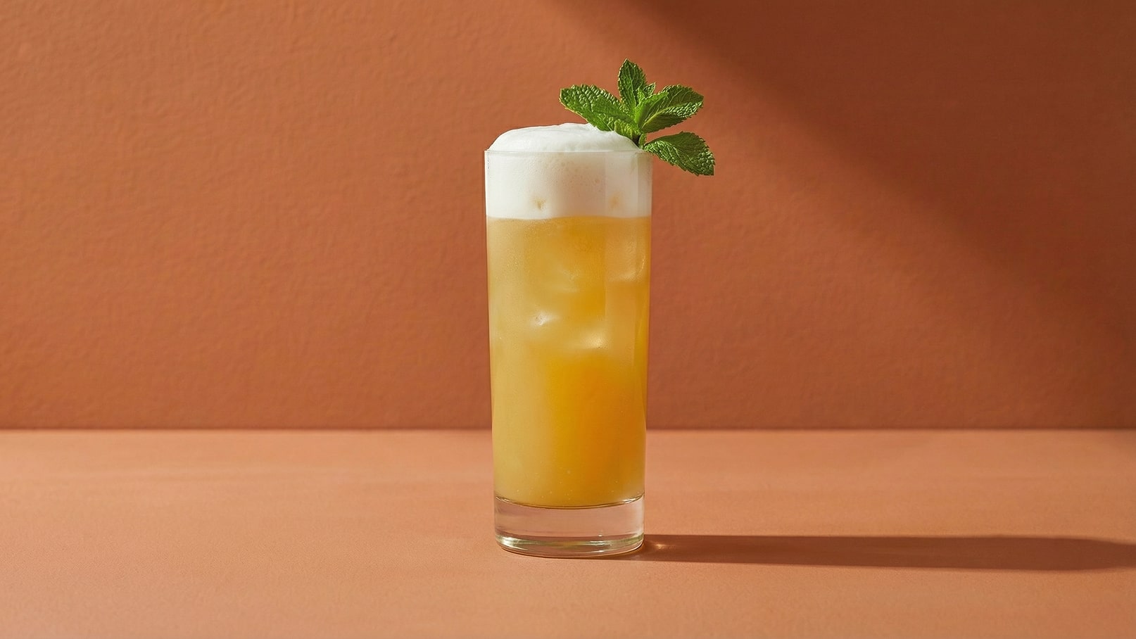 Ein hoher Glasbecher enthält ein gelbliches Getränk mit Eiswürfeln. Obenauf befindet sich eine schaumige Schicht und ein frisches Minzblatt. Der Hintergrund ist in einem warmen Orangeton gehalten.