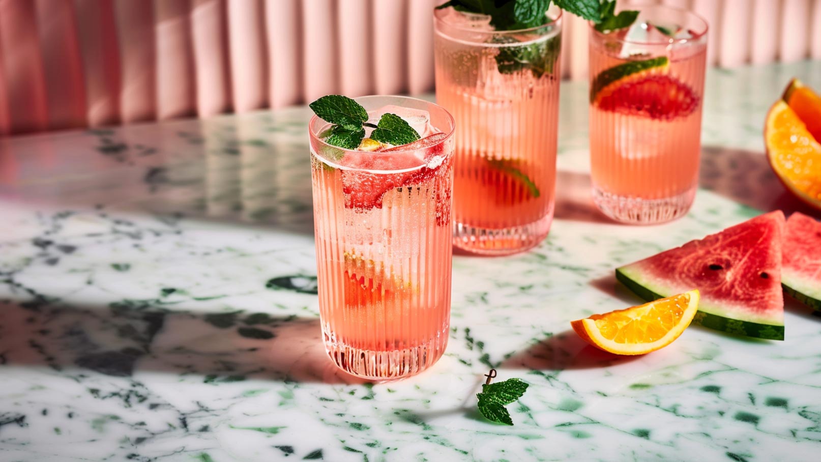 Ein bunt gemixter Cocktail in einem festen Glas, gefüllt mit Eis und einer orangefarbenen Scheibe. Im Hintergrund sind Wassermelonen und Stücke von Wassermelone sichtbar. Der Cocktail hat einen fruchtigen Look und scheint erfrischend zu sein.