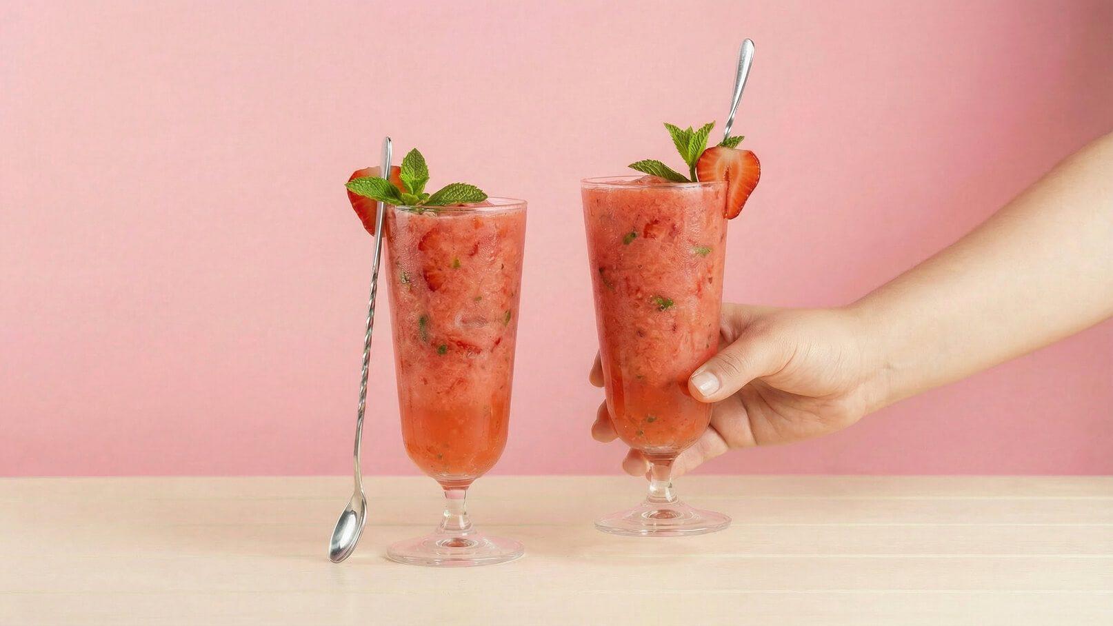 Zwei Gläser mit einem fruchtigen, gefrorenen Getränk aus Erdbeeren und Minze stehen auf einem Tisch. Ein Hand greift nach dem rechten Glas, während das andere Glas mit einem Löffel dekoriert ist. Der Hintergrund ist rosa.