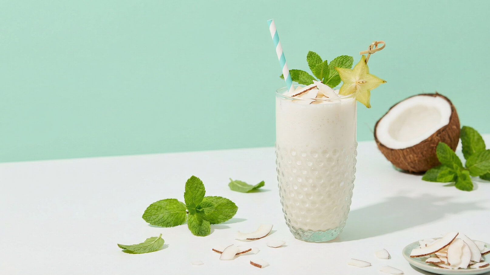 Ein erfrischendes Getränk in einem transparenten Glas mit einem gestreiften Trinkhalm. Der Smoothie ist mit Kokosraspeln und einem Stern aus Frucht dekoriert. Grünes Minzblatt liegt daneben, und auf dem Tisch sind Kokosnussstückchen verteilt. Der Hintergrund ist mintgrün.