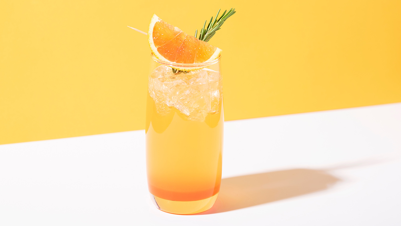 Auf dem Bild ist ein Glas mit einem erfrischenden Passionsfrucht-Orangen-Cocktail zu sehen. Zur Dekoration sind eine Blutorangenscheibe, Eiswürfel und ein Rosmarinzweig im Glas platziert. Die leuchtenden Farben des Cocktails und die frischen Zutaten verleihen dem Bild eine sommerliche und ansprechende Optik. Der Rosmarinzweig setzt einen aromatischen Akzent, der die Sinne anspricht. 