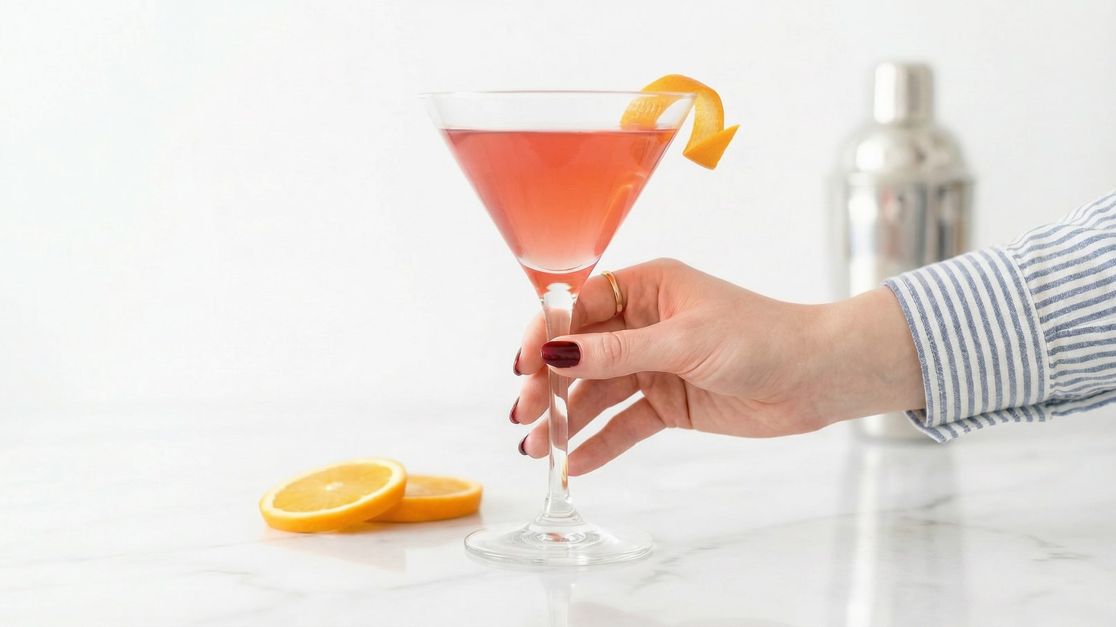 Ein Glas mit einem pinkfarbenen Getränk, dekoriert mit einer Orangenscheibe, wird von einer Hand gehalten. Im Hintergrund liegt eine geschnittene Orangenscheibe auf einer Marmortheke.