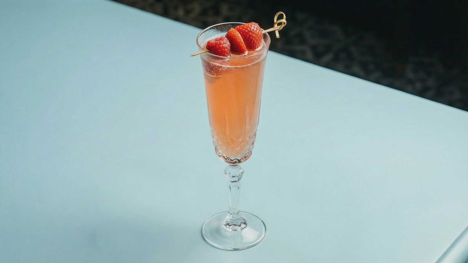 Ein hohes, elegantes Glas mit einem sprudelnden, rosafarbenen Getränk. Oben sind drei frische Erdbeeren auf einem Zahnstocher dekoriert. Der Hintergrund ist unscharf und zeigt eine helle Fläche.