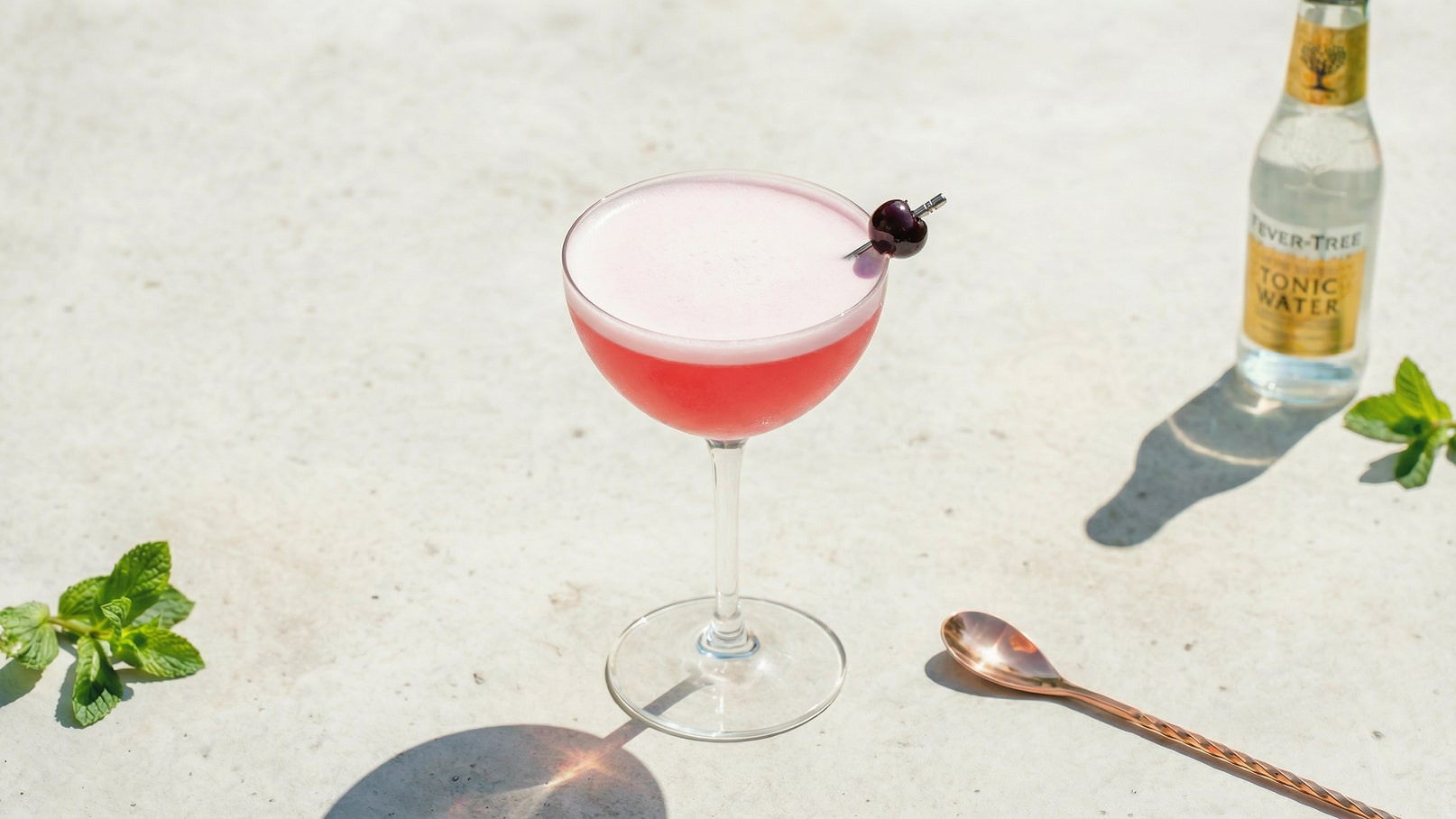 Ein elegantes Cocktailglas mit einem pinkfarbenen Drink und einer Schaumkrone. Eine Kirsche steckt auf einem kleinen Spieß im Glas. Im Hintergrund sind frische Minzblätter und eine Flasche Tonic Water zu sehen.