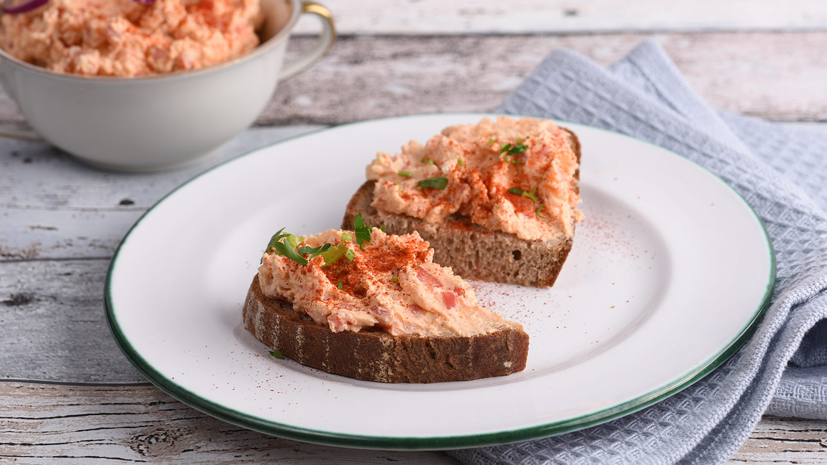 Auf einem weißen Teller sind zwei Scheiben Brot mit Liptauer-Aufstrich und frischen Kräutern appetitlich angerichtet. Im Hintergrund steht eine Tasse mit Liptauer. Der Teller steht auf einem blauen Tuch, das eine frische Atmosphäre schafft. Einladend und ansprechend für Liebhaber von herzhaften Vorspeisen.