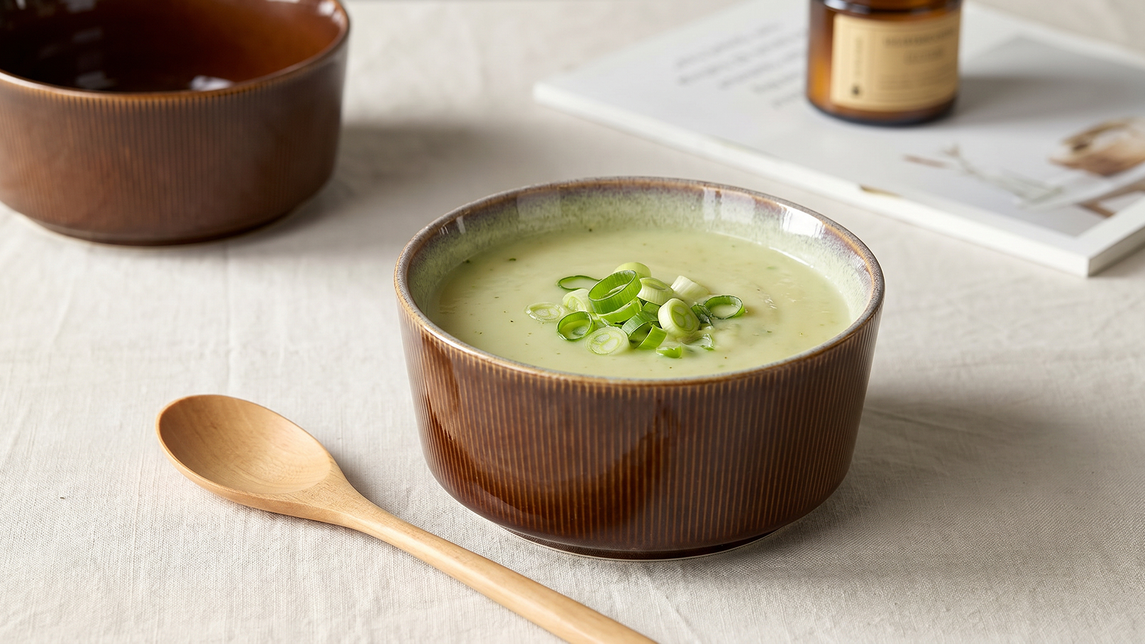 Eine braune Schüssel mit cremiger, grüner Suppe, garniert mit gehackten Frühlingszwiebeln. Daneben liegt ein Holzlöffel. Im Hintergrund sind ein weiterer Teller und ein Glas zu sehen. Die Umgebung ist hell und klar.