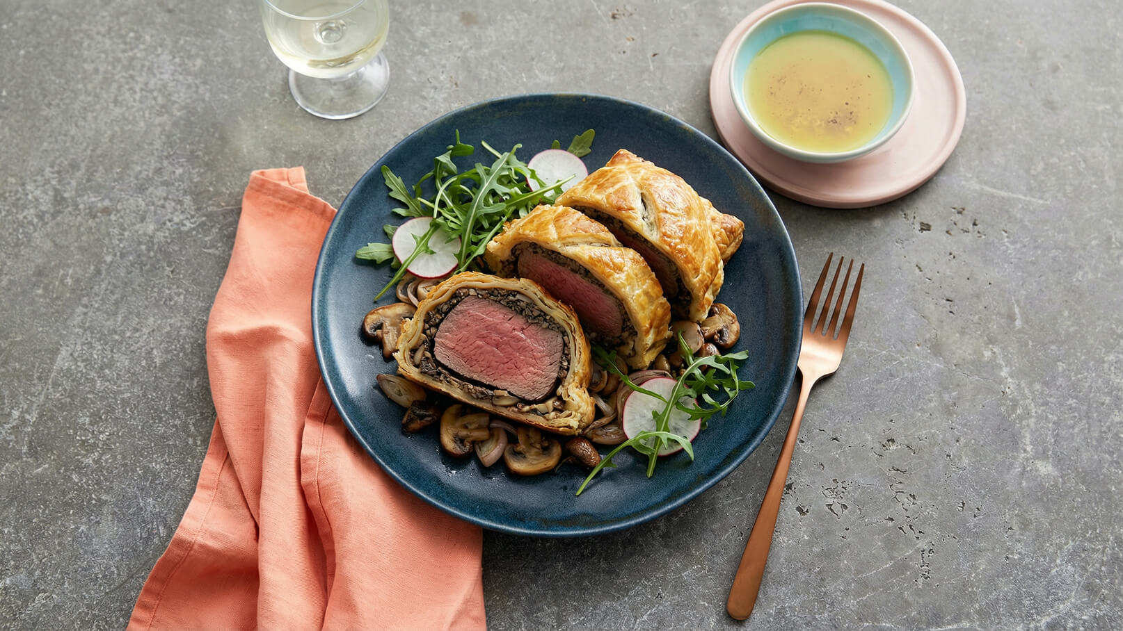 Auf dem Bild ist ein Teller mit einer portionierten Beef Wellington zu sehen, umgeben von gebratenen Pilzen, frischem Rucola und Radieschen. Neben dem Teller liegt eine orangefarbene Serviette und ein Glas mit Weißwein steht im Hintergrund.