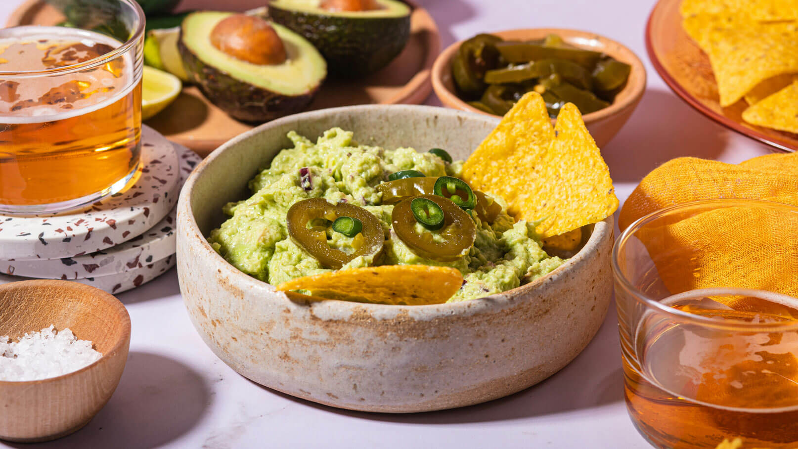 Das Bild zeigt eine elegante schwarze Schüssel, randvoll gefüllt mit cremiger Guacamole und großzügig garniert mit frischen Kräutern. Die intensive grüne Farbe der Guacamole sticht hervor und lädt zum Probieren ein. Um die Schüssel herum liegen knusprige Nachos, bereit zum Dippen in die leckere Guacamole. Zudem liegen frische Limetten, die für eine erfrischende Note sorgen.