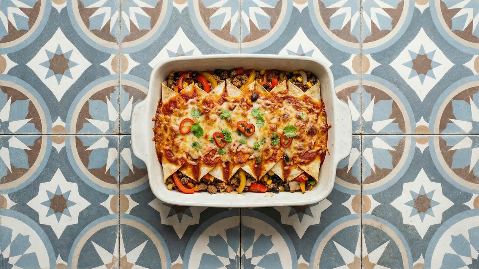 Eine Auflaufform mit Enchiladas, gefüllt mit Fleisch, buntem Gemüse und einer Schicht geschmolzenem Käse. Die Enchiladas sind mit frischen Jalapeños und Koriander garniert, auf einem gemusterten Fliesenboden platziert.