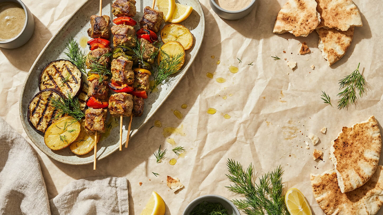 Ein ovaler Teller mit gegrillten Fleischspießen, umgeben von Zitrone, gegrilltem Gemüse und frischen Kräutern. Daneben ein Schälchen mit einer Dipsauce und ein Stück Fladenbrot, das auf einem braunen, gewellten Untergrund liegt.