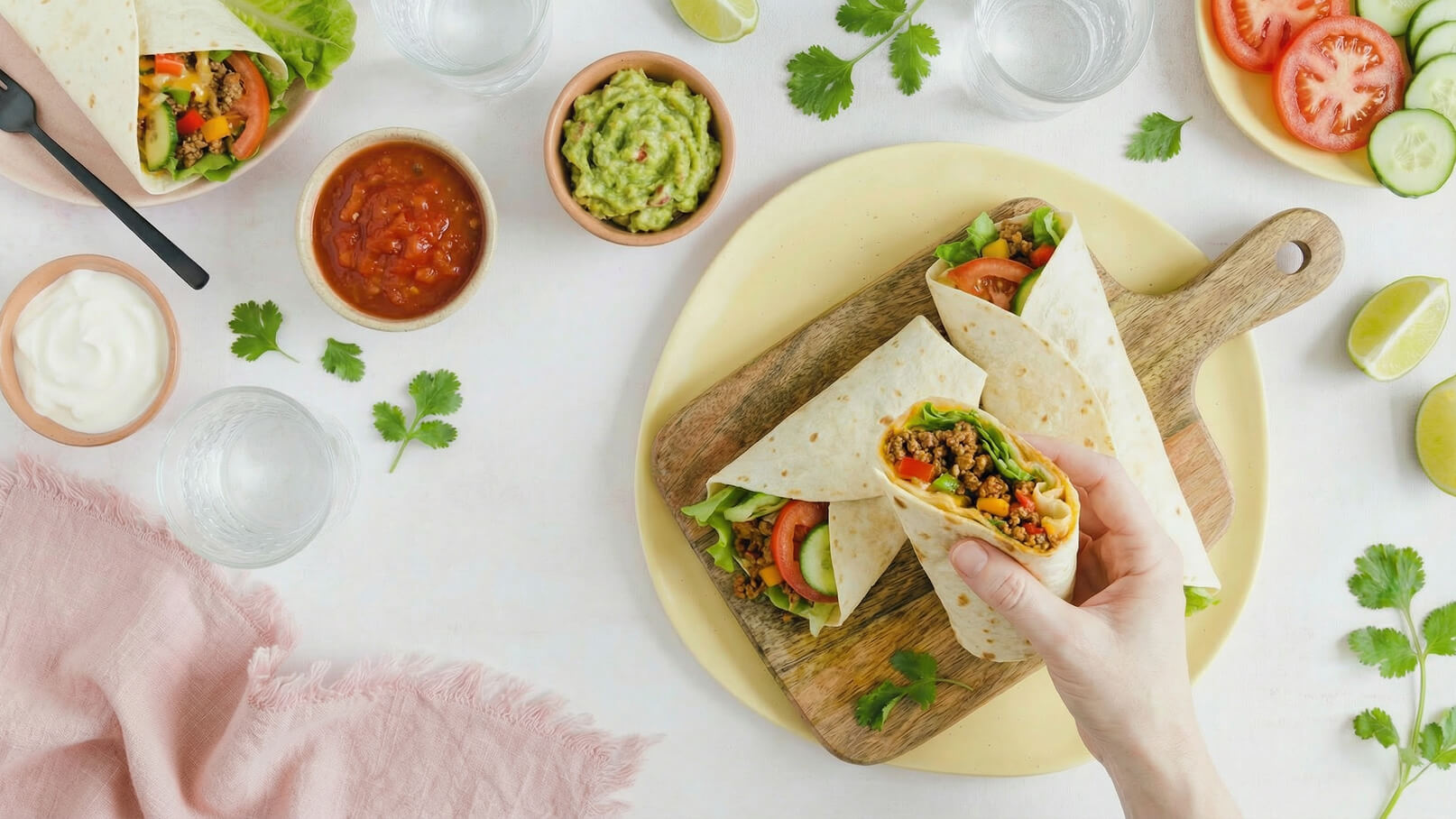 Eine Hand hält einen gefüllten Wrap mit Gemüse, während zwei weitere Wraps auf einem Holzbrett liegen. Daneben befinden sich Schüsseln mit Guacamole und Salsa, sowie eine halbe Limette und frische Kräuter. Wasser und frisches Gemüse sind ebenfalls im Bild zu sehen.