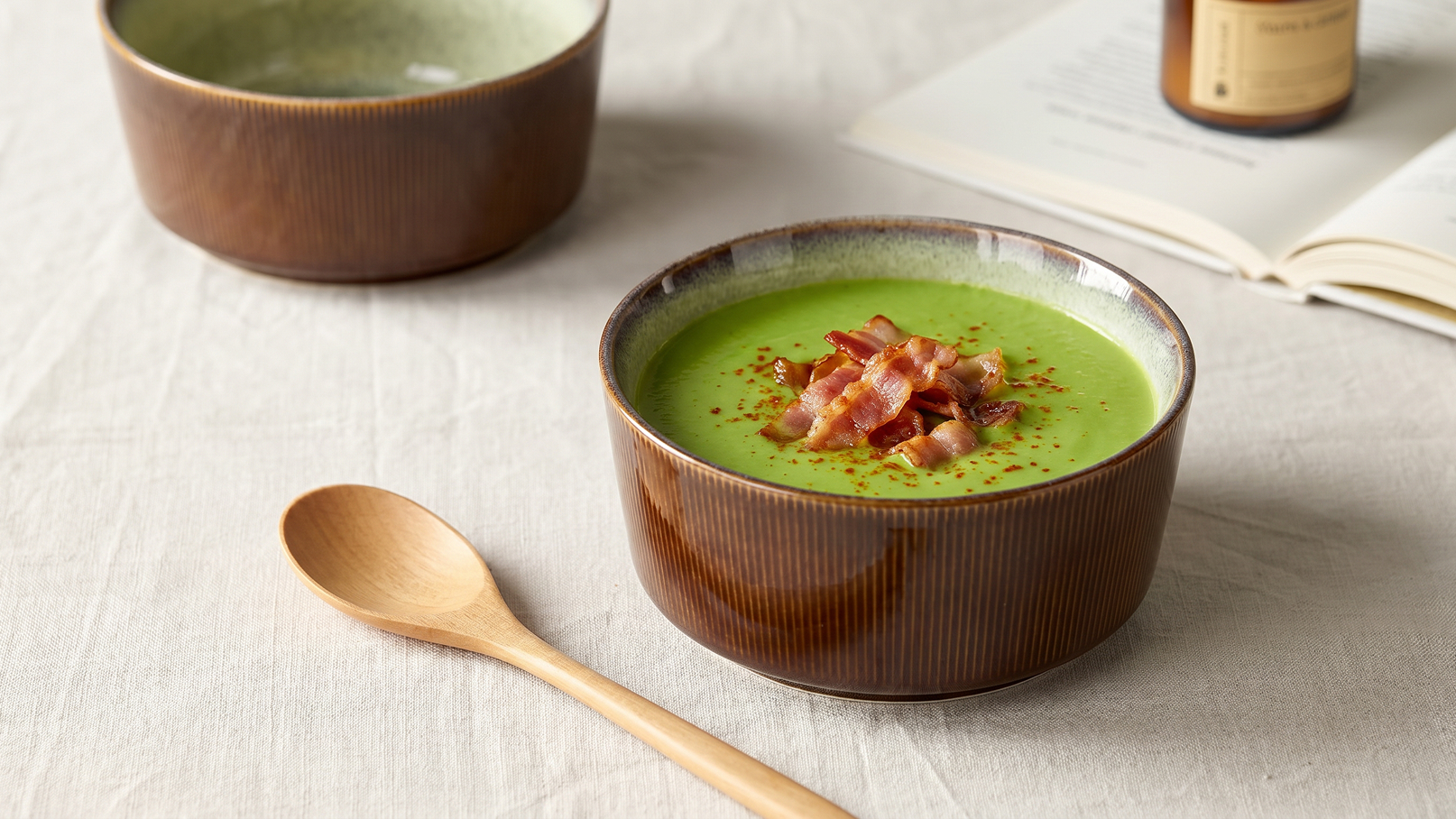 Eine grüne Suppe in einer braunen Schüssel, garniert mit knusprigem Speck und roten Gewürzen. Neben der Schüssel liegt ein Holzlöffel. Im Hintergrund sind weitere Schüsseln und ein aufgeschlagenes Buch zu sehen.