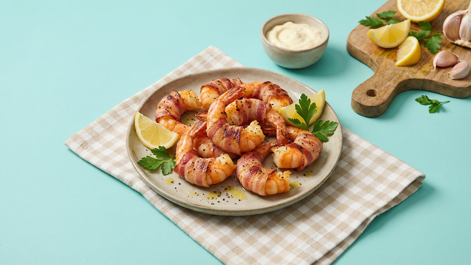 Ein Teller mit gegrillten Garnelen, umwickelt mit Speck, wird auf einem karierten Tuch präsentiert. Garniert mit grüner Petersilie und Zitronenscheiben. Im Hintergrund befindet sich eine kleine Schüssel mit Sauce und einige Knoblauchzehen. Der Hintergrund ist in sanftem Türkis gehalten.