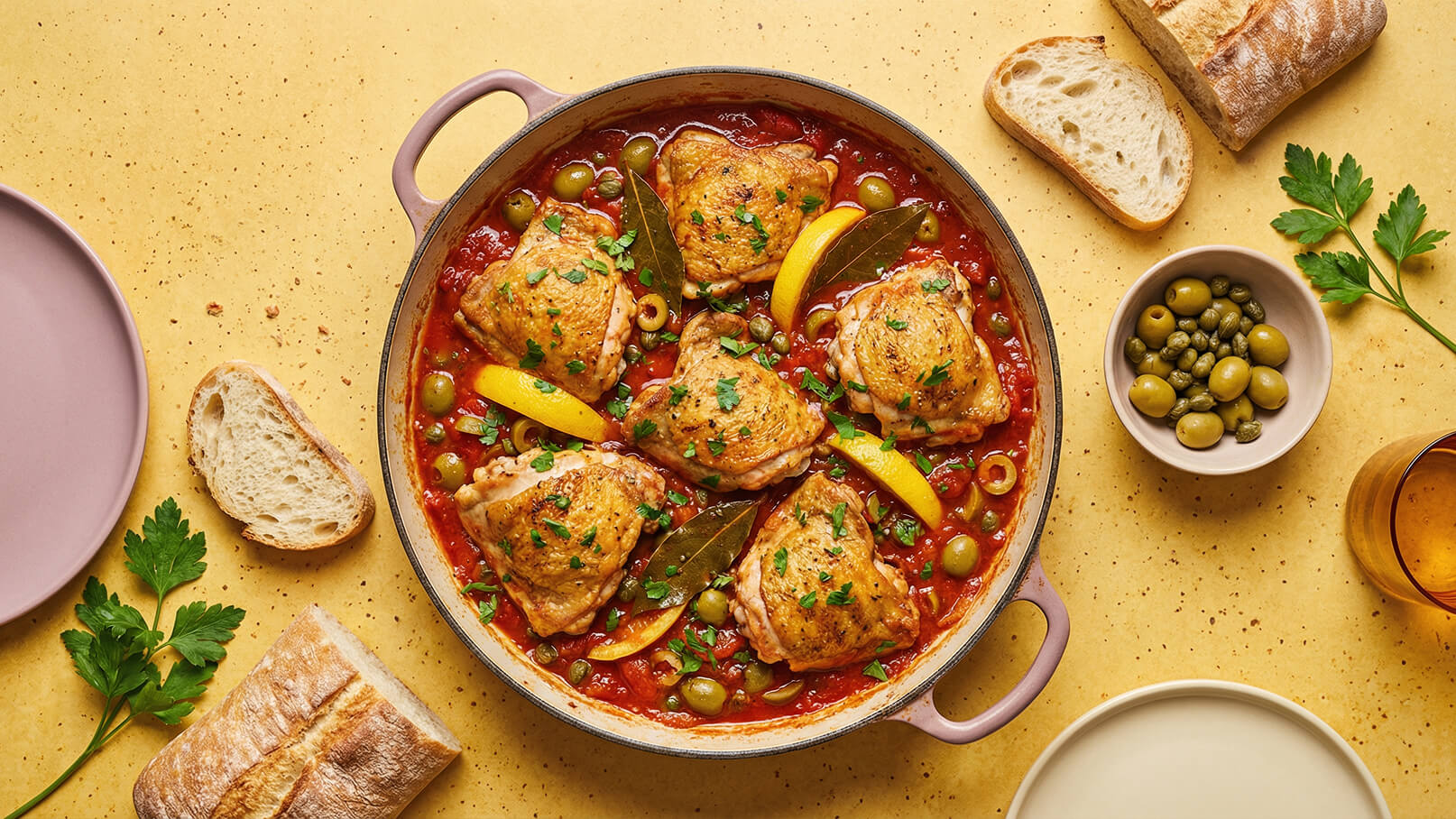 Ein großer Topf mit saftigen Hähnchenschenkeln, die in einer tomatigen Soße mit grünen Oliven und Zitronenscheiben gekocht werden. Frische Petersilie ist darüber gestreut. Daneben liegen Stücke von Brot auf einer gelben Tischfläche.