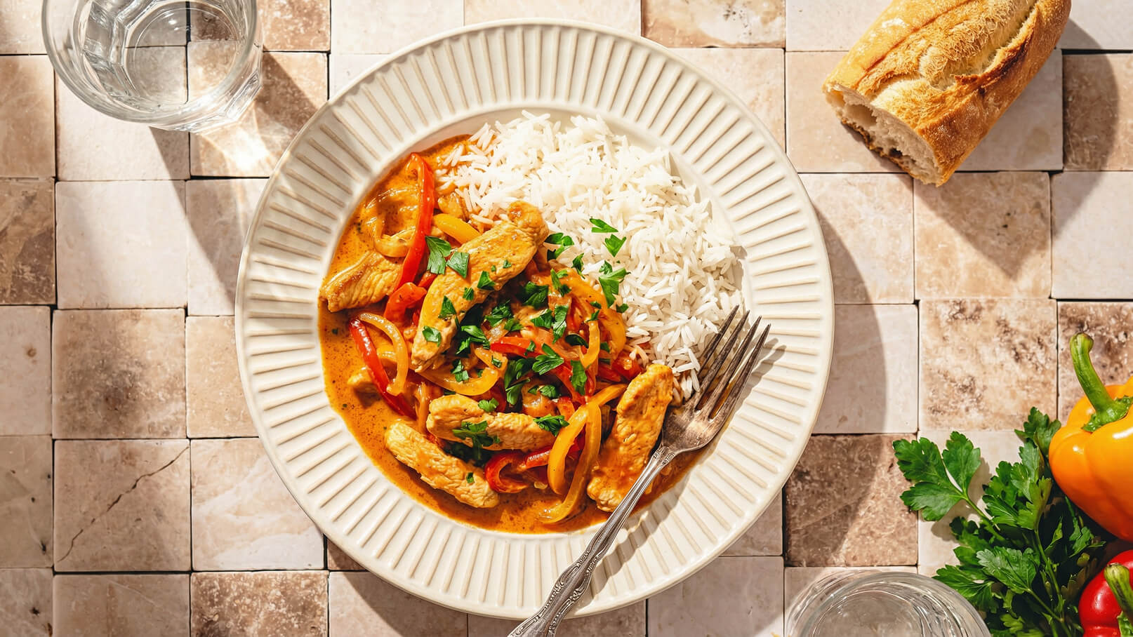 Ein Teller mit Hühnchen in einer bunten Paprika-Sauce, serviert mit weißem Reis. Das Gericht ist mit frischer Petersilie garniert. Daneben liegt ein Besteck und ein Glas Wasser. Im Hintergrund sind frische Paprika und Kräuter sichtbar.
