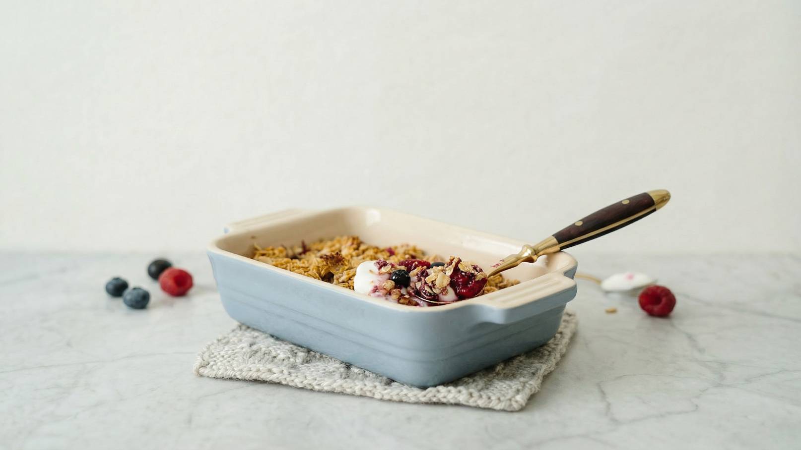 Eine graue Auflaufform mit einer Mischung aus Müsli, Joghurt und Früchten. Ein goldfarbener Löffel mit einem dunklen Griff liegt in der Form. Daneben befinden sich einige frische Heidelbeeren und Himbeeren auf einem hellen Untergrund.