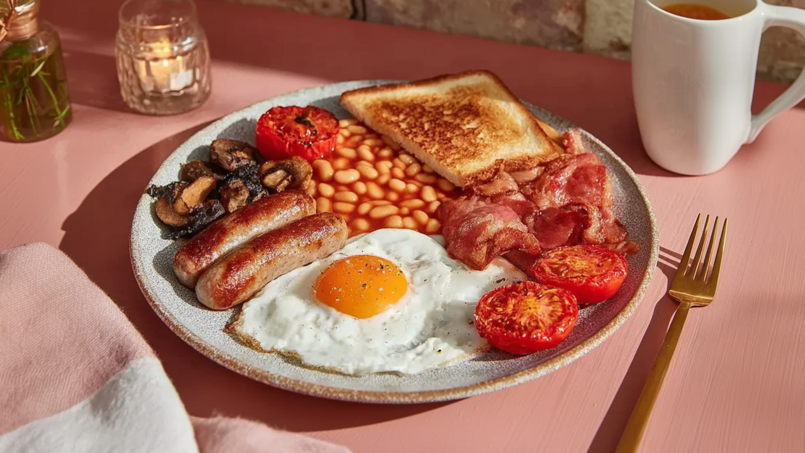Ein Frühstücksteller mit Spiegelei, gebratenem Speck, Würstchen, gegrillten Tomaten, gebackenen Bohnen und gebuttertem Toast. Neben dem Teller steht eine Tasse. Der Tisch ist in sanften Farben dekoriert.